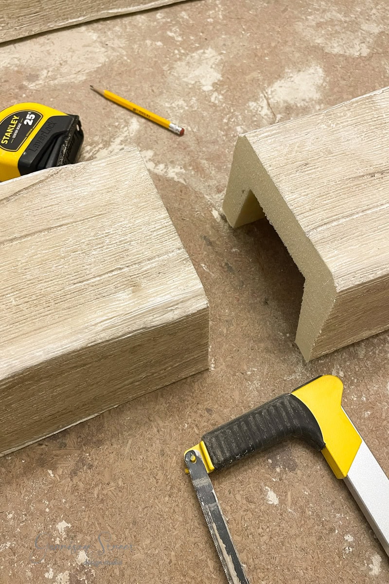 Two pieces of wood with U-shaped cuts lie on a workbench. A yellow tape measure, a pencil, and a hacksaw are nearby.