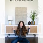 A person with long, curly hair sits cross-legged in front of a white IKEA Kallax sideboard with fluted doors, pointing at it. The sideboard is topped with a lamp, plant, and decorative panel. Text reads "DIY Ikea Kallax Hack Makeover with Fluted Doors.