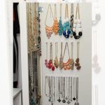 A white wall-mounted jewelry organizer displays necklaces and earrings of various colors and styles. The text reads "DIY Ikea Nissedal Wall Mirror Hidden Jewelry Hack" by Garrison Street Design Studio.