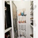 A narrow closet space featuring a DIY IKEA NISSEDAL wall mirror jewelry holder. Colorful necklaces and earrings are neatly displayed behind the the mirror with shelves on either side storing clothes and shoes. Text: "DIY IKEA NISSEDAL WALL MIRROR HIDDEN JEWELRY HACK, garrison street design studio.