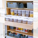 A neatly organized pantry shelf with clear labeled containers holding grains and pasta, along with small baskets storing canned goods and dry items. The space is efficiently utilized for easy access and visibility.