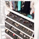 An organized walk-in closet with IKEA Pax features hanging clothes, multiple sunglasses, and jewelry neatly arranged in pull out trays.
