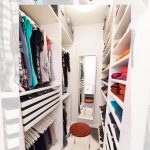 A modern, organized walk-in closet with white shelving, colorful clothes neatly folded and hanging, a narrow mirror on the wall, and a small stool with a red cushion in the center. Ambient lighting illuminates the space.