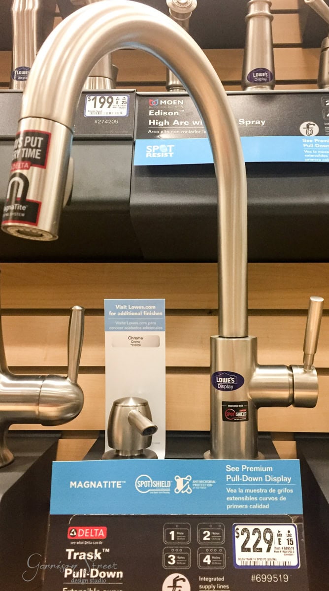 A display of a stainless steel pull-down kitchen faucet with a curved spout. Price tag shows $229 at a hardware store. Various features and labels like "SpotShield" and "MagnaTite" are visible on the tags.