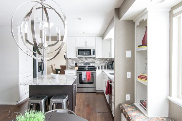 Modern kitchen with white cabinets, stainless steel appliances, and a marble island. A geometric chandelier hangs above the island, which has three metal stools. Light streams through windows, and a built-in shelf displays decor items.