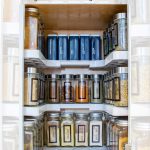 Image of a well-organized spice cabinet with labeled glass jars and metal containers neatly arranged on shelves. The text above reads, "Easy Kitchen Spice Cabinet Storage Organization - Garrison Street Design Studio.