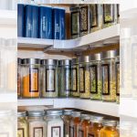Organized spice cabinet with labeled jars and metal containers neatly arranged on shelves. Contains various spices and herbs in transparent and metal containers. Text overlay reads "Easy Kitchen Spice Cabinet Storage Organization.