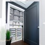 A neatly organized linen closet with black and white patterned bins on top shelves, neatly folded towels in the middle, and IKEA Trofast storage units with labeled drawers below. The closet door is black and partially open.