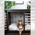 A small dog sits cozily on a pillow inside a DIY dog crate made from an Ikea Malm dresser. The crate features decorative tiles in the back and a wooden slat design on the side. Green plants adorn the top of the dresser. Text above reads: "Modern DIY Ikea Malm Dog Crate Hack from a Dresser, garrison street design studio.