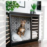 A cute brown dog sits comfortably inside a DIY dog crate made from a modified IKEA Malm dresser. The crate features a cozy pillow, black and white patterned walls, and an open design. A plant and decor sit on top of the furniture piece.