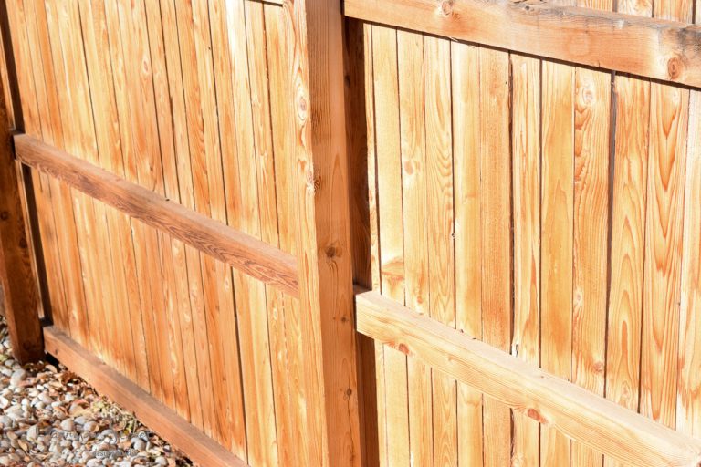 A wooden fence with a vertical plank design, showing natural wood grain and texture. Sunlight casts shadows, highlighting the warm tones. Pebbles are visible at the base of the fence.