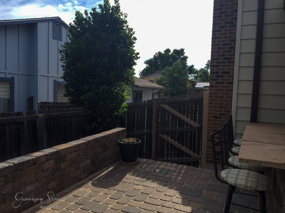 A patio area with brick flooring and a small wall, next to a tall brick and siding house. There's a potted plant and three cushioned stools near a wooden fence gate. Trees and neighboring houses are visible in the background.