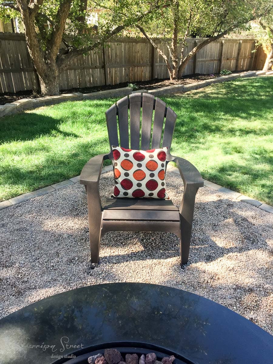 A brown Adirondack chair with a colorful polka dot pillow sits on a gravel area in a backyard with green grass and a wooden fence. Trees provide shade in the background.