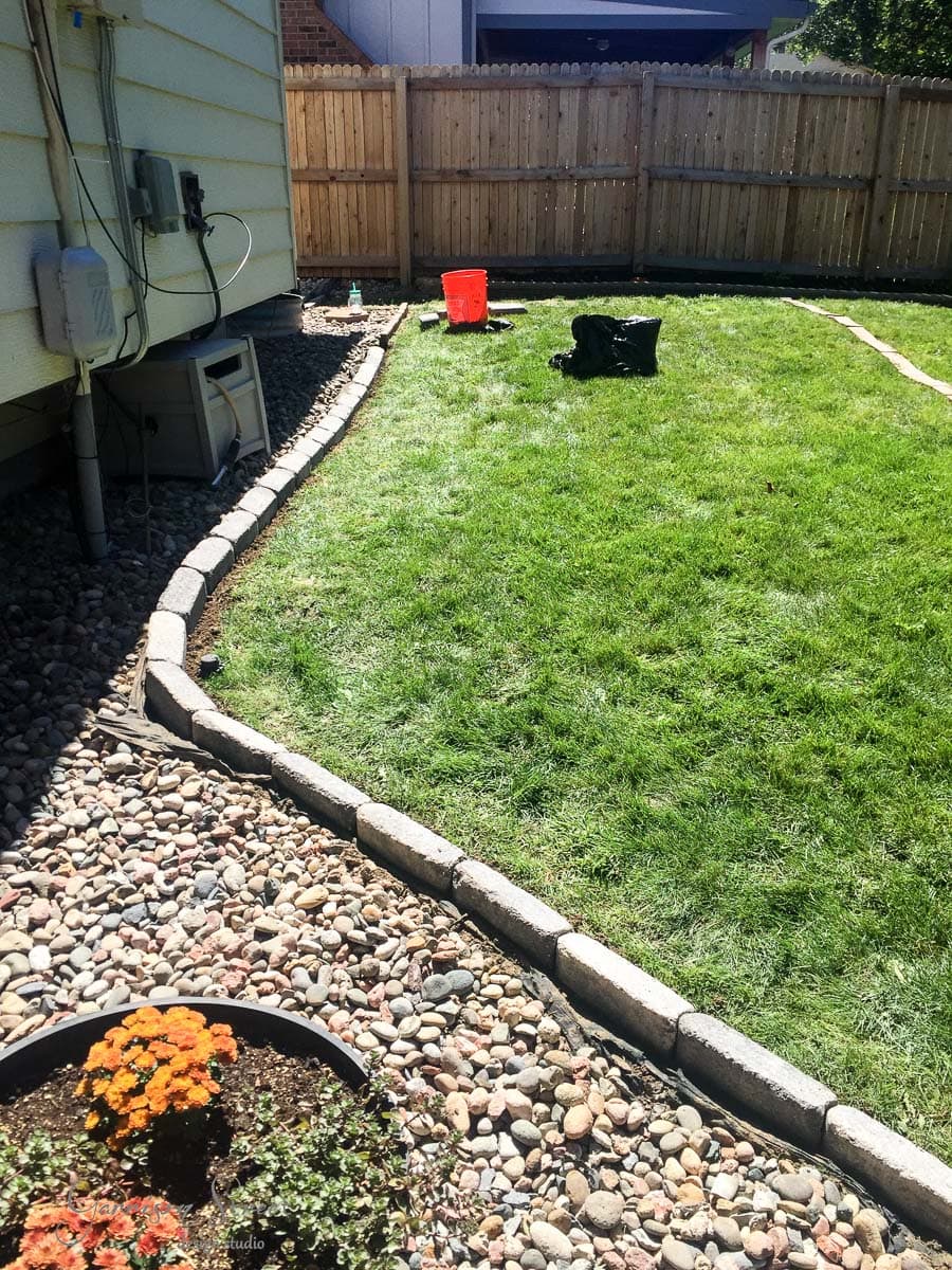 A sunny backyard with a green lawn bordered by rocks. A black backpack and a red bucket are on the grass. A small garden with orange flowers is visible in the foreground, near the house with cream siding. A wooden fence encloses the yard.