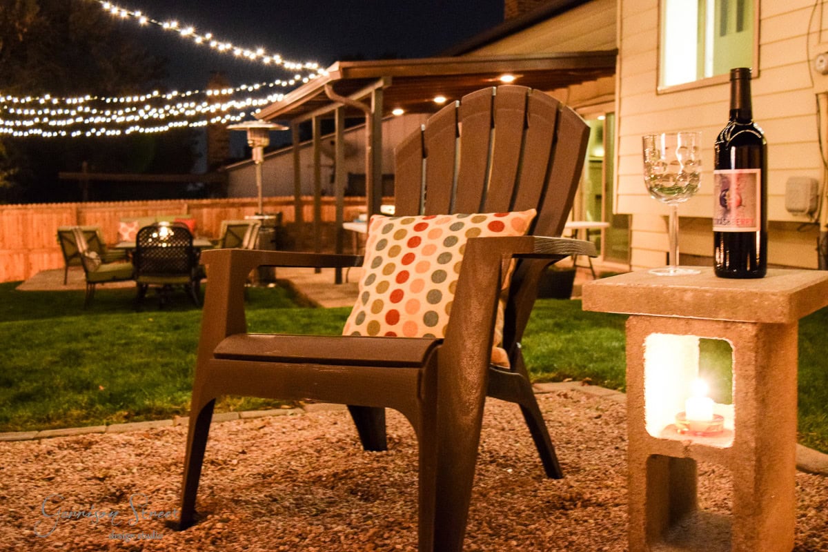 A cozy backyard at night featuring a large chair with a colorful polka dot pillow, a bottle of wine, and two glasses on a concrete side table. String lights and a seating area with a fire pit add warmth to the scene.