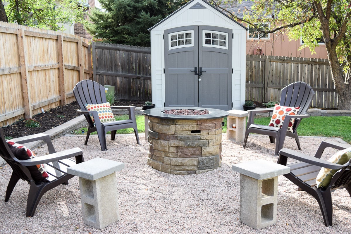 A backyard features a circular stone fire pit surrounded by four Adirondack chairs with colorful cushions. The area is covered in gravel, with concrete blocks as side tables. A small gray and white shed is visible in the background, enclosed by a wooden fence.