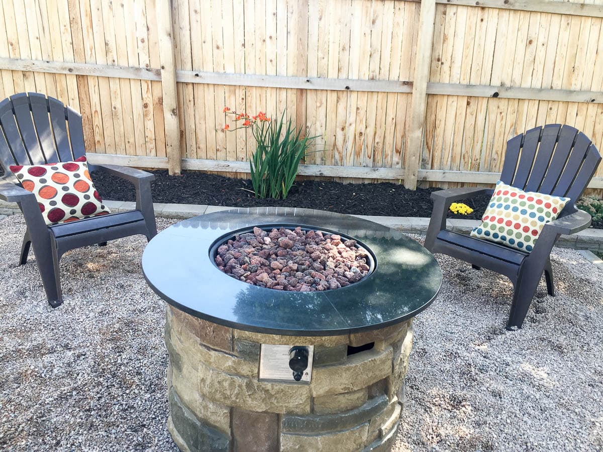Outdoor seating area with two Adirondack chairs, each with a colorful patterned cushion. A stone fire pit filled with rocks is in between. A wooden fence and plants, including orange flowers, are in the background, and the ground is covered with gravel.
