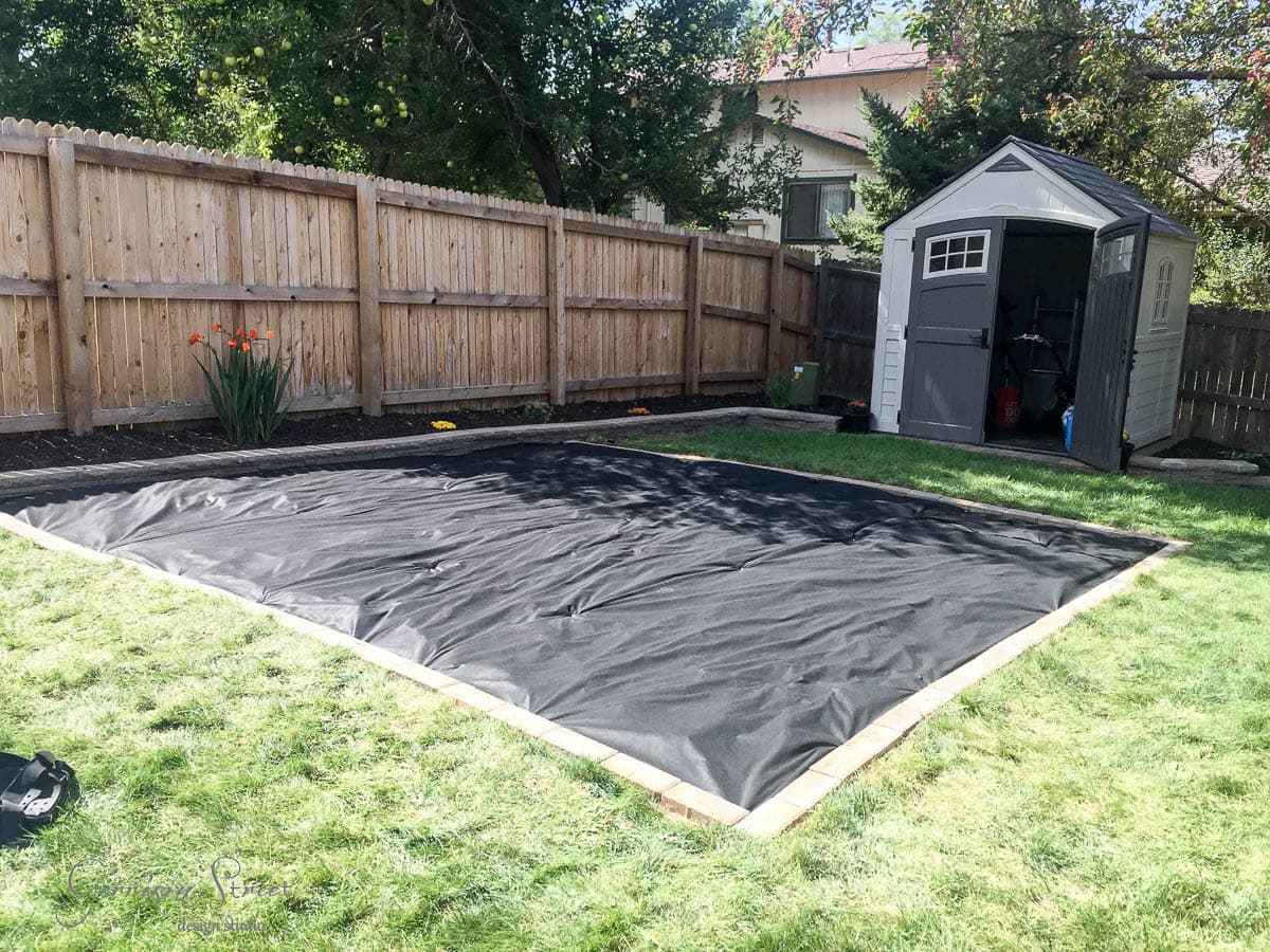 A backyard with weed barrier laid out on the grass, bordered by bricks. A wooden fence surrounds the yard, and a small shed is in the corner. Bright orange flowers grow near the fence.