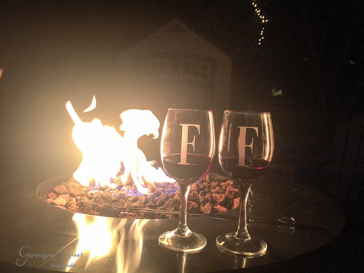 Two wine glasses with the letter "F" are placed on a table near a glowing fire pit. The scene is set against a dark evening backdrop with a softly lit building and string lights visible in the distance.