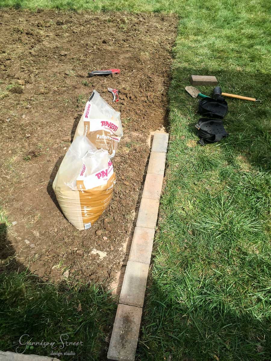 A garden area with a row of bricks separating soil from grass. Two bags of paver sand are on the soil. Nearby, a red-handle tool, black knee pads, and a shovel are on the ground.