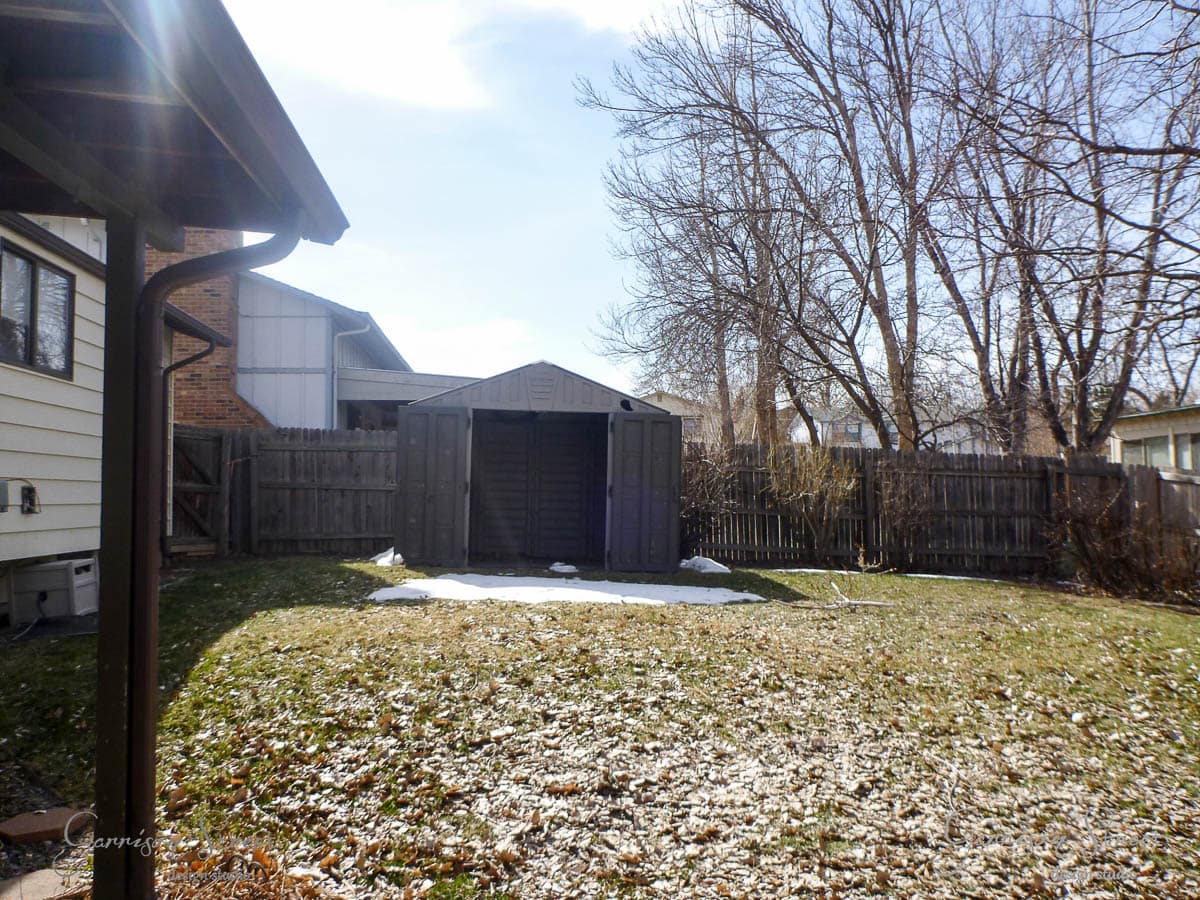 A backyard with a brown storage shed centered against a wooden fence. The ground is partially covered with snow and dry grass. Bare trees and neighboring houses are visible under a blue sky with some clouds.