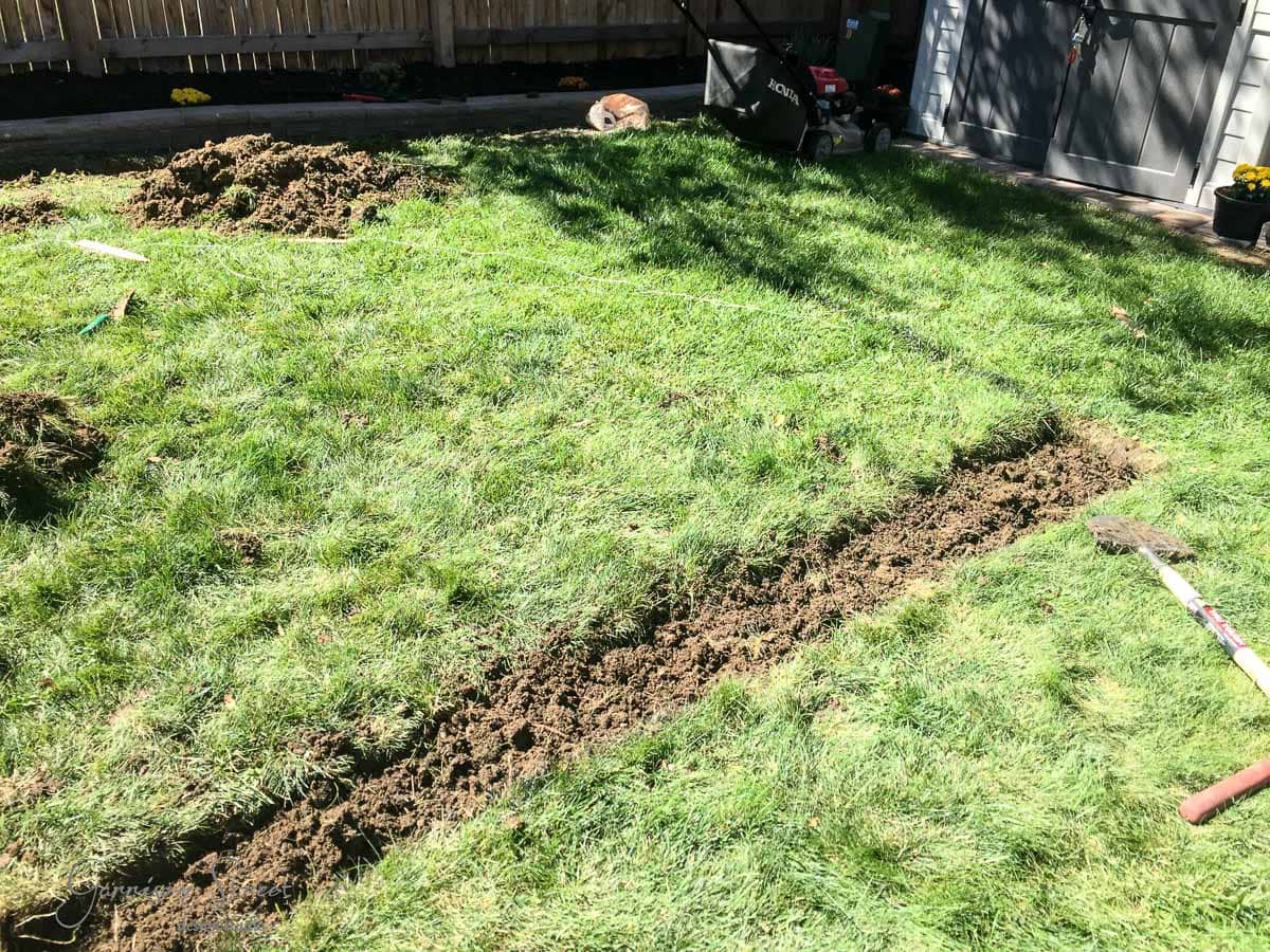 A grassy backyard being prepared for landscaping. A trench runs diagonally through the grass, with soil piled on the side. Garden tools and a lawn mower are visible. A wooden fence and a small shed are in the background.