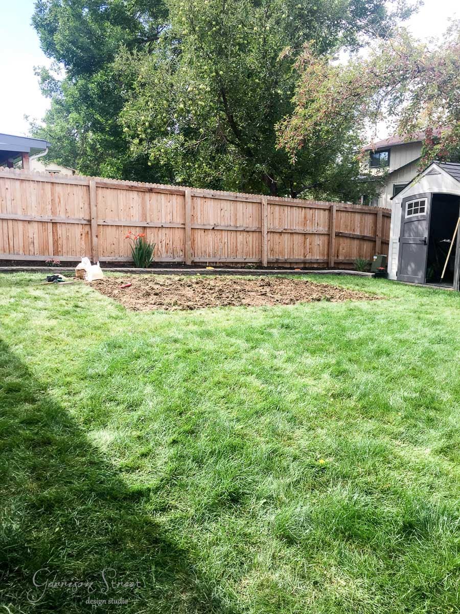 A backyard garden with a freshly tilled rectangular patch of soil. A wooden fence borders the yard, and there is a small shed on the right. A few trees are in the background.