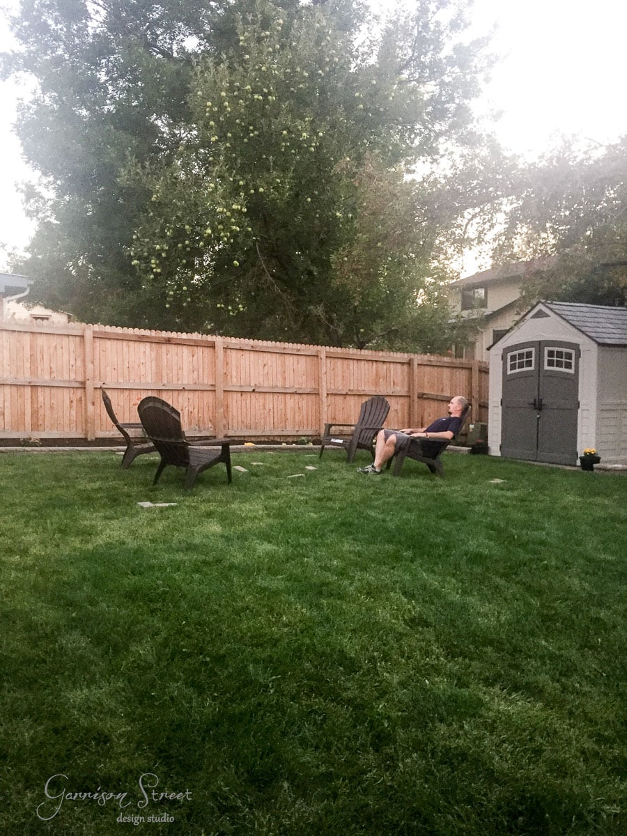 A man is sitting on an outdoor chair in a grassy backyard surrounded by a wooden fence. There are several empty chairs nearby, and a small shed is in the corner under large trees. The sky is partially visible with hints of a sunset.