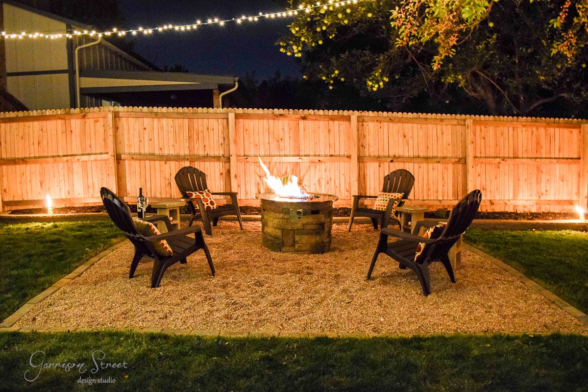 A cozy backyard scene at night, featuring a stone fire pit surrounded by four Adirondack chairs on a gravel surface. String lights hang overhead, and a wooden fence encloses the area. A bottle of wine and glasses rest on the ground near a chair.