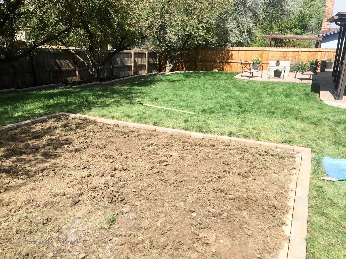 A backyard with a rectangular patch of tilled soil, bordered by bricks. The rest of the yard is grassy with trees and a wooden fence. In the distance, there is a patio with furniture and potted plants.
