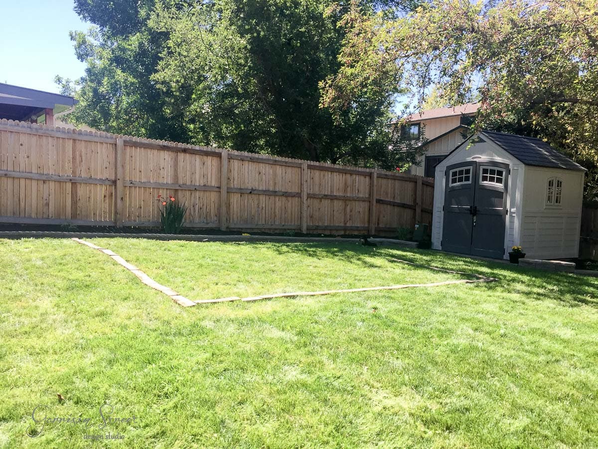A sunny backyard with a green lawn and a wooden fence, featuring a small garden shed with a dark roof in the corner. There is a simple stone path creating a rectangle on the grass. Trees provide some shade along the fence line.