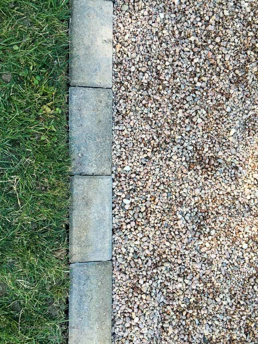 A top view of a garden path featuring a border of rectangular bricks. On one side, there is green grass, and on the other side, pea gravel.