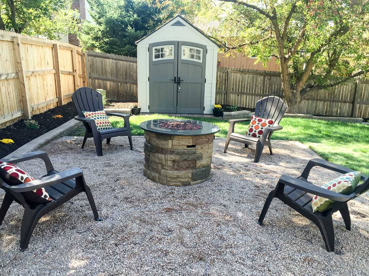 A backyard scene featuring a stone fire pit surrounded by four black Adirondack chairs with colorful patterned cushions. The area is covered in gravel and bordered by a wooden fence and green grass. A small shed is in the background under a tree.