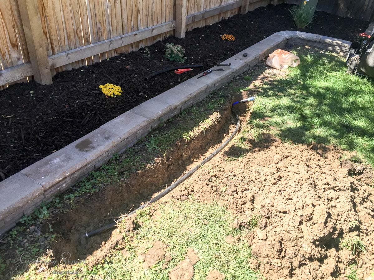 A garden scene with a freshly dug trench containing a black irrigation pipe along a brick-bordered flower bed. Soil is piled next to the trench, and garden tools and a bag are visible. A wooden fence and greenery are in the background.