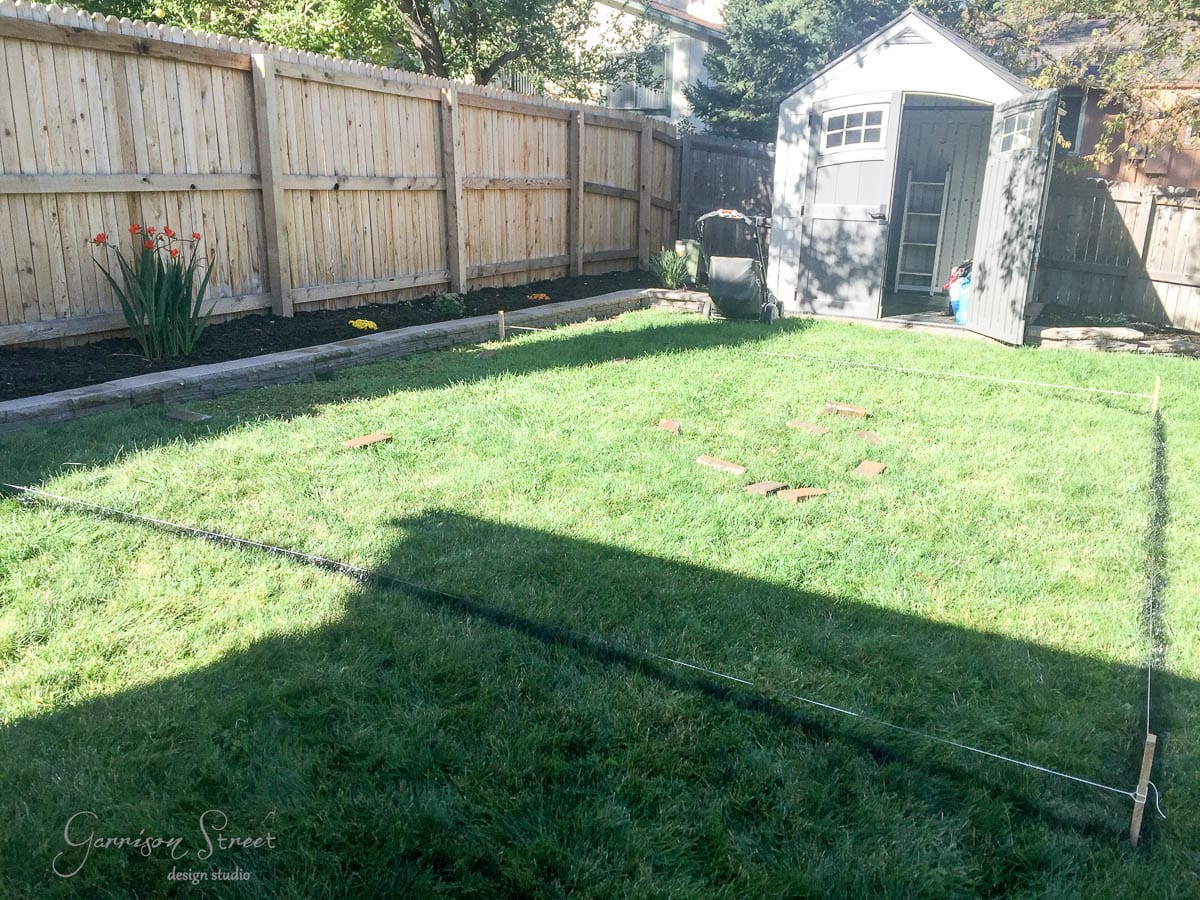 A sunny backyard with a section of grass marked off by black string and stakes. Red bricks are scattered within the marked area. A wooden fence surrounds the yard, and a shed is in the corner. Tulips bloom along the fence.