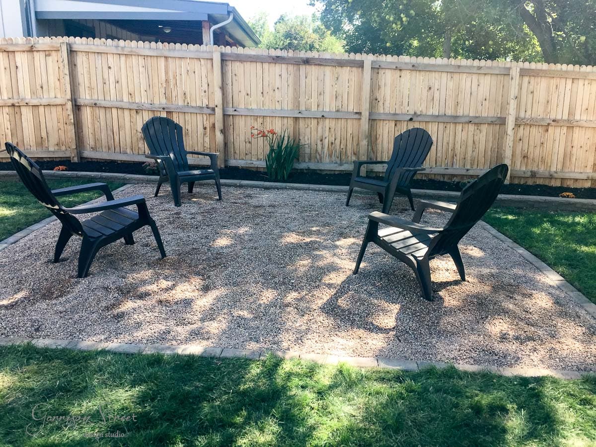 A small outdoor seating area features four black Adirondack chairs arranged in a circle on a gravel surface. The area is surrounded by a wooden fence and bordered by a narrow strip of grass. A few plants grow near the fence.