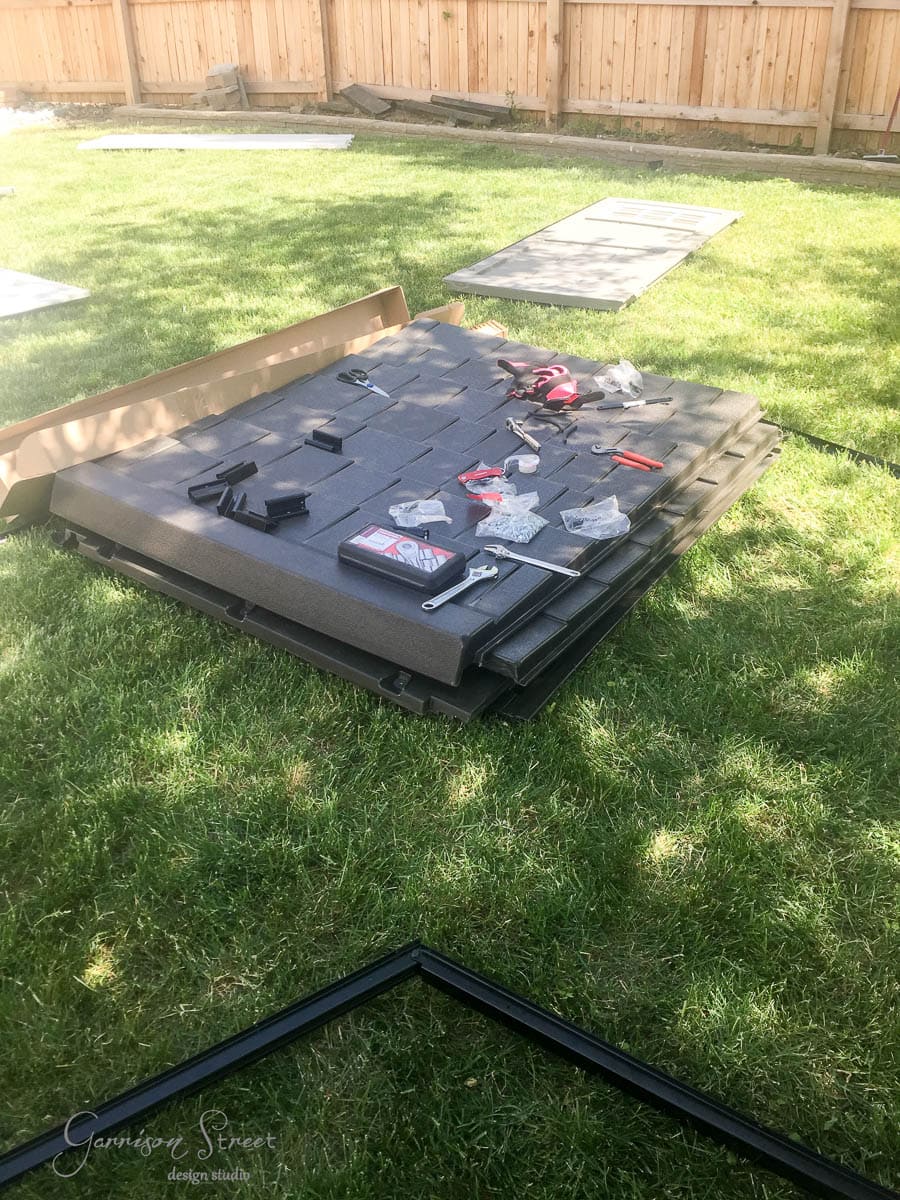 Metal roof panels are laid out on grass alongside tools and hardware for assembly. A wooden fence surrounds the yard in the background, and parts of a frame are visible.