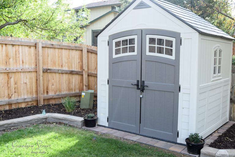 A small white and gray garden shed with double doors and arched windows stands on a stone path. It's situated in a backyard with a wooden fence, green lawn, and a couple of potted plants nearby.