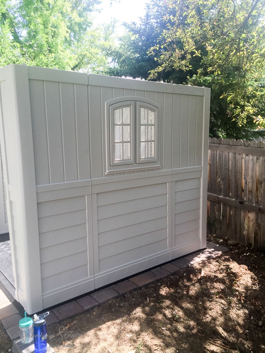 A small, white garden shed with a window stands on a paved area in a backyard. It is surrounded by a wooden fence and trees, casting light shadows. Two spray cans are placed at the base of the shed.