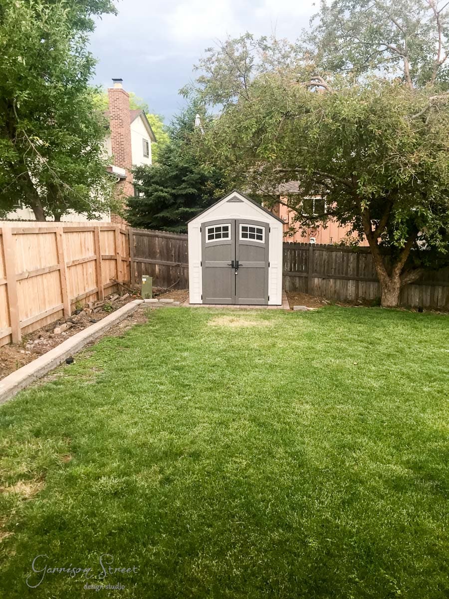 A small gray and white shed stands at the end of a neatly maintained grassy backyard. It's surrounded by trees and bordered by a wooden fence on the left. A brick house sits in the background.