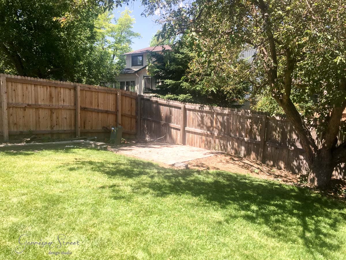 A sunny backyard with a wooden fence surrounds a grassy area. A tree with shade is on the right, and a small patio with paving stones is at the center. A house is partially visible in the background.