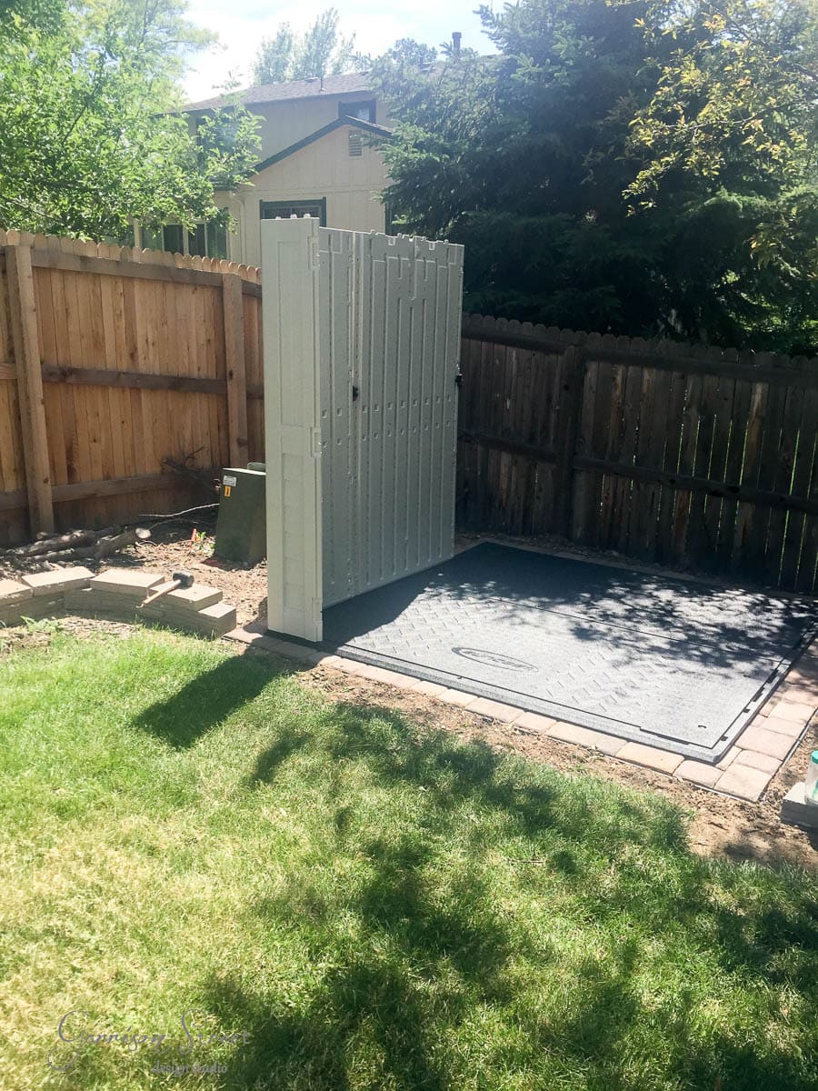 A small outdoor shower stall with a gray base and light gray walls is set up in a grassy backyard. It is positioned near a wooden fence, with trees providing natural shade.