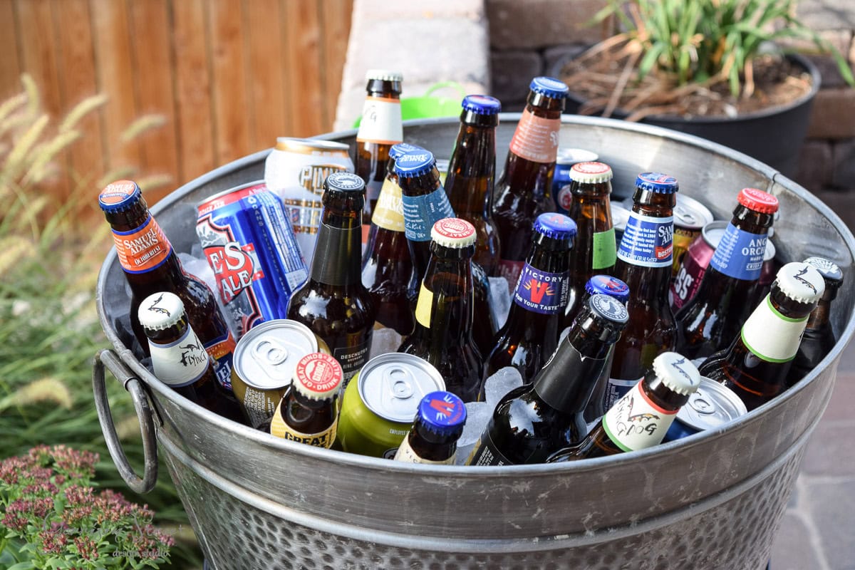 A large metal tub filled with a variety of beer and soda bottles, each with colorful caps and labels, surrounded by ice. The tub is placed outdoors near plants and a wooden fence.