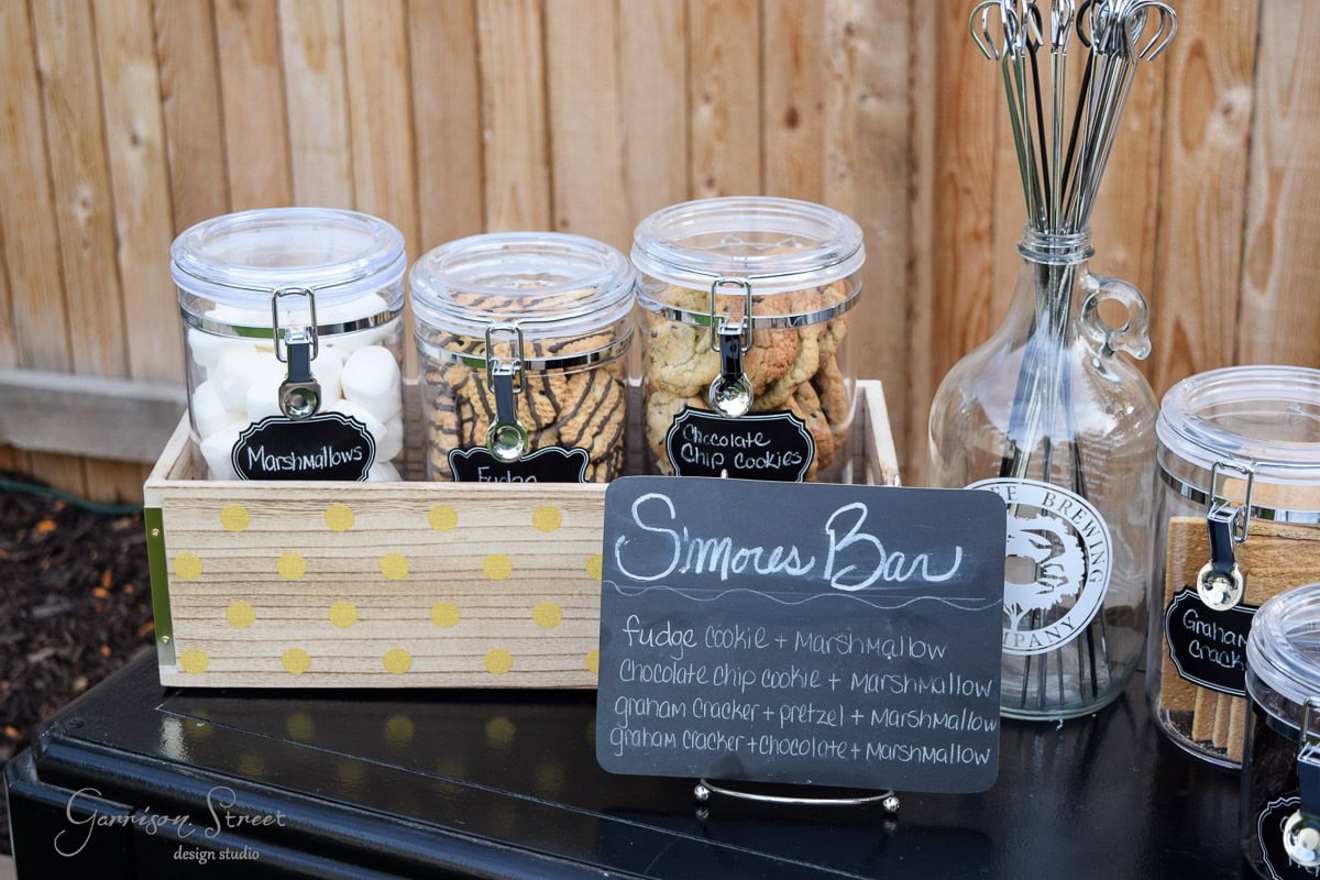 A wooden box with jars labeled "Marshmallows," "Fudge," and "Chocolate Chip Cookies" on a black table. A sign reads "S'mores Bar." Nearby stands a bottle holding skewers. A wooden fence serves as the background.