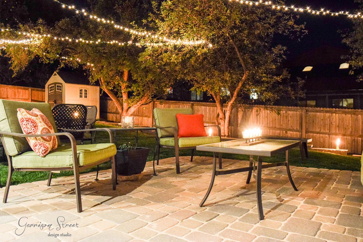 Cozy backyard patio at night with string lights overhead. Green cushioned seating surrounds a glass-top table with lit candles. A wooden fence and trees are in the background, along with a small, lit-up garden shed.