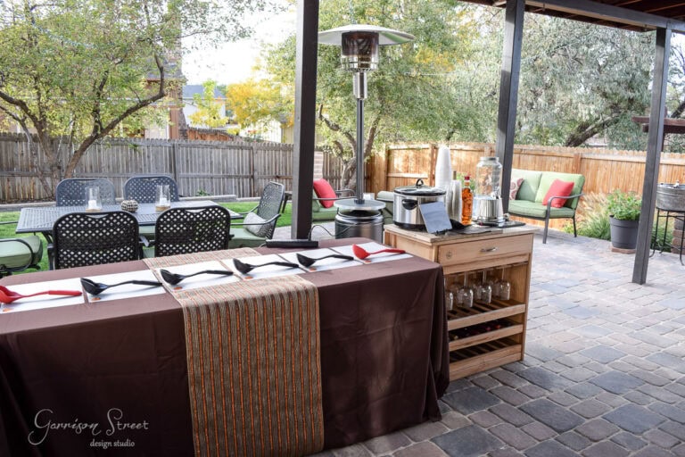 Outdoor patio set up for a gathering, featuring a table with brown cloth and table runner, neatly arranged cutlery and napkins, another table with drink dispensers and snacks, surrounded by cushioned seating, and trees in the background.