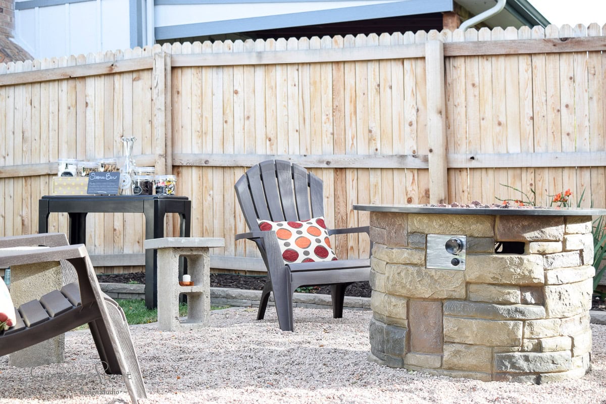 Outdoor patio with a wooden fence, Adirondack chairs, a side table, and a brick fire pit. A black table holds party supplies in the background. The seating area is adorned with a colorful polka dot cushion on one chair.