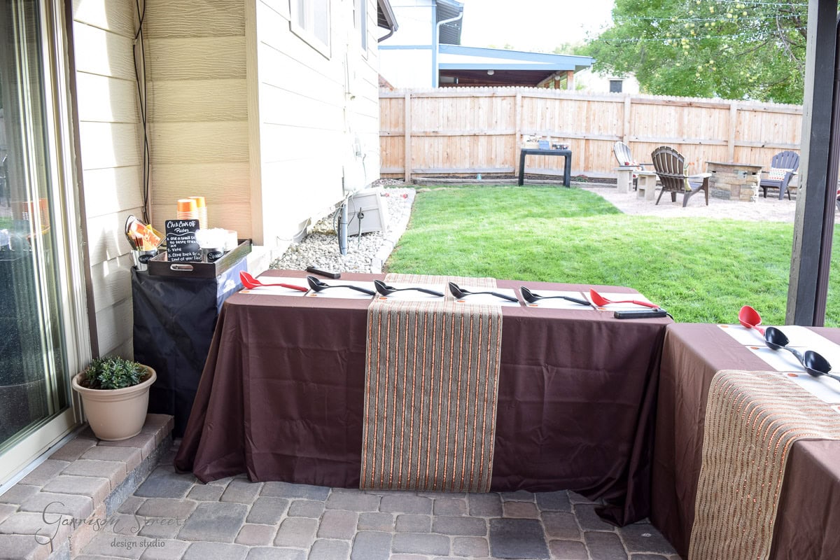 Outdoor patio with tables set for a gathering. Brown tablecloths and striped runners adorn the tables, which hold utensils and napkins. A small plant sits beside a drink station. A grassy yard and chairs around a fire pit are visible in the background.
