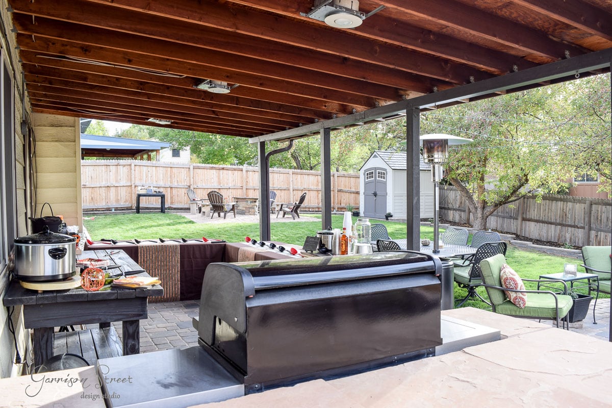 Covered patio with a wooden roof, grill, and outdoor seating area. A sofa and chairs with green cushions face a coffee table. The backyard features a lawn, fire pit with chairs, and a storage shed against a wooden fence.