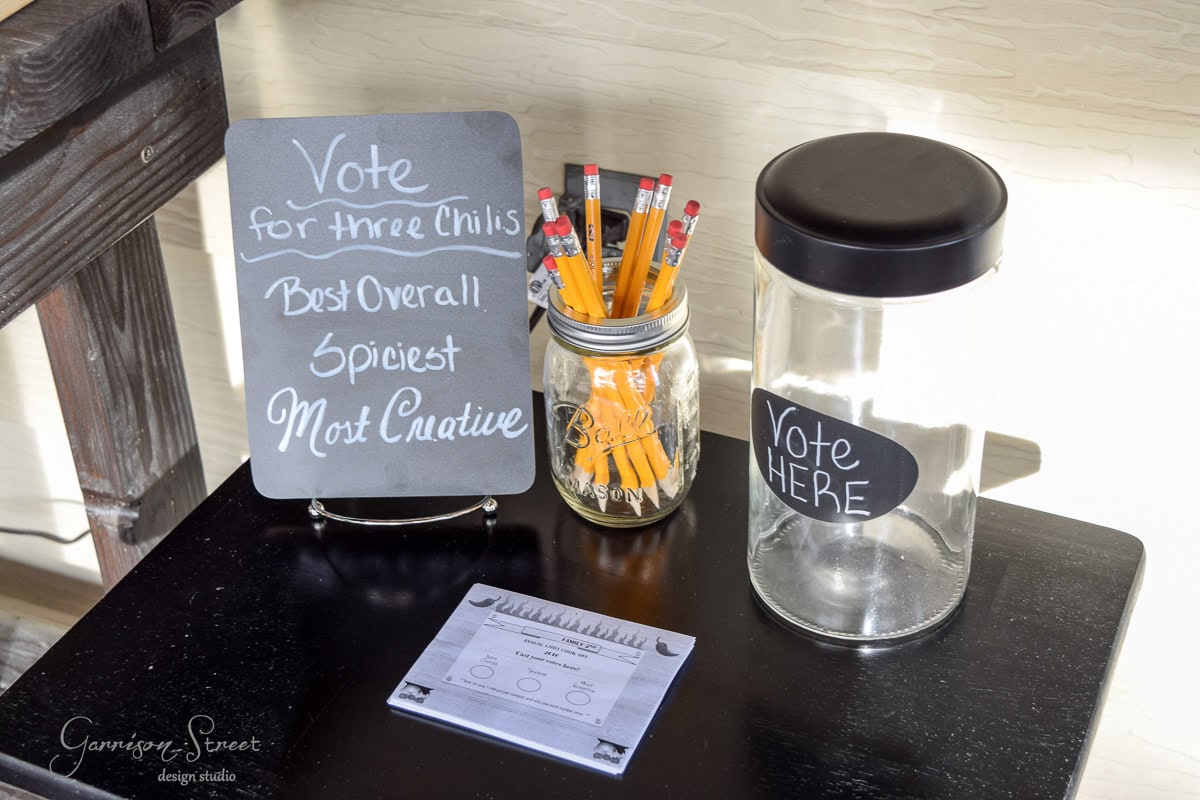 A small table with a chalkboard sign reads "Vote for three chilis: Best Overall, Spiciest, Most Creative." A jar of pencils, a blank voting sheet, and a glass jar labeled "Vote Here" are placed beside it.